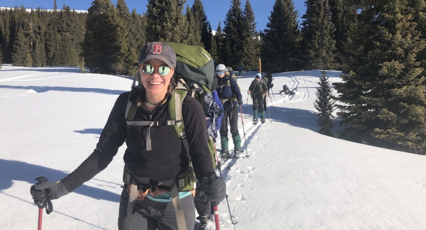 A person wearing winter gear and a backpack smiles. They are in front of other students, walking behind.